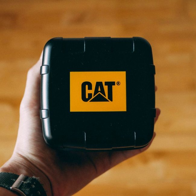 Close-up of a hand holding a black box with CAT branding over a wooden floor. Perfect for industrial and product themes.