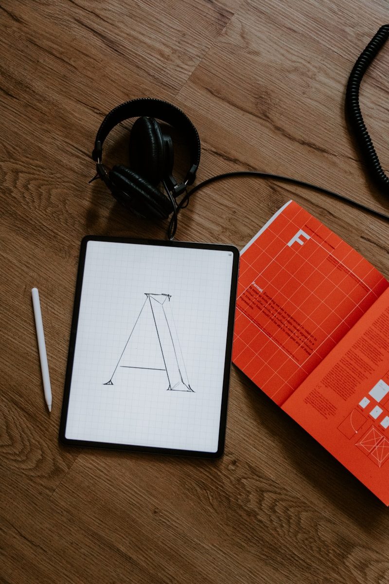A tablet with a letter A sketch, stylus, headphones, and orange book on wood